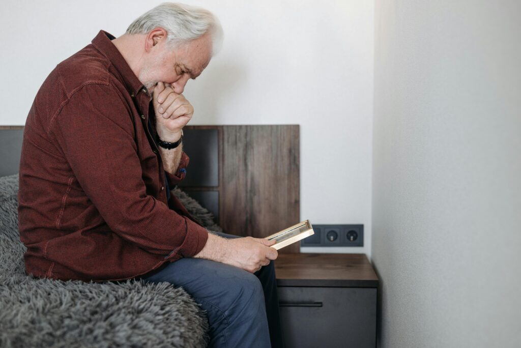 Senior man sitting indoors, holding a photo, reflecting on memories and experiencing sorrow.