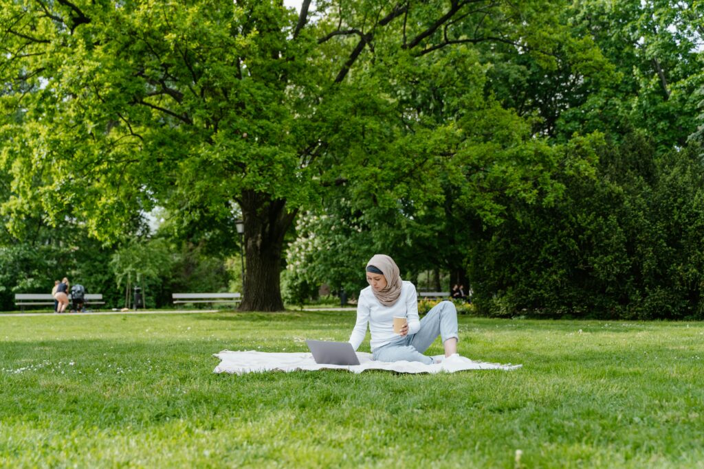 A Muslim woman in a hijab working on her laptop in a vibrant green park, enjoying nature.